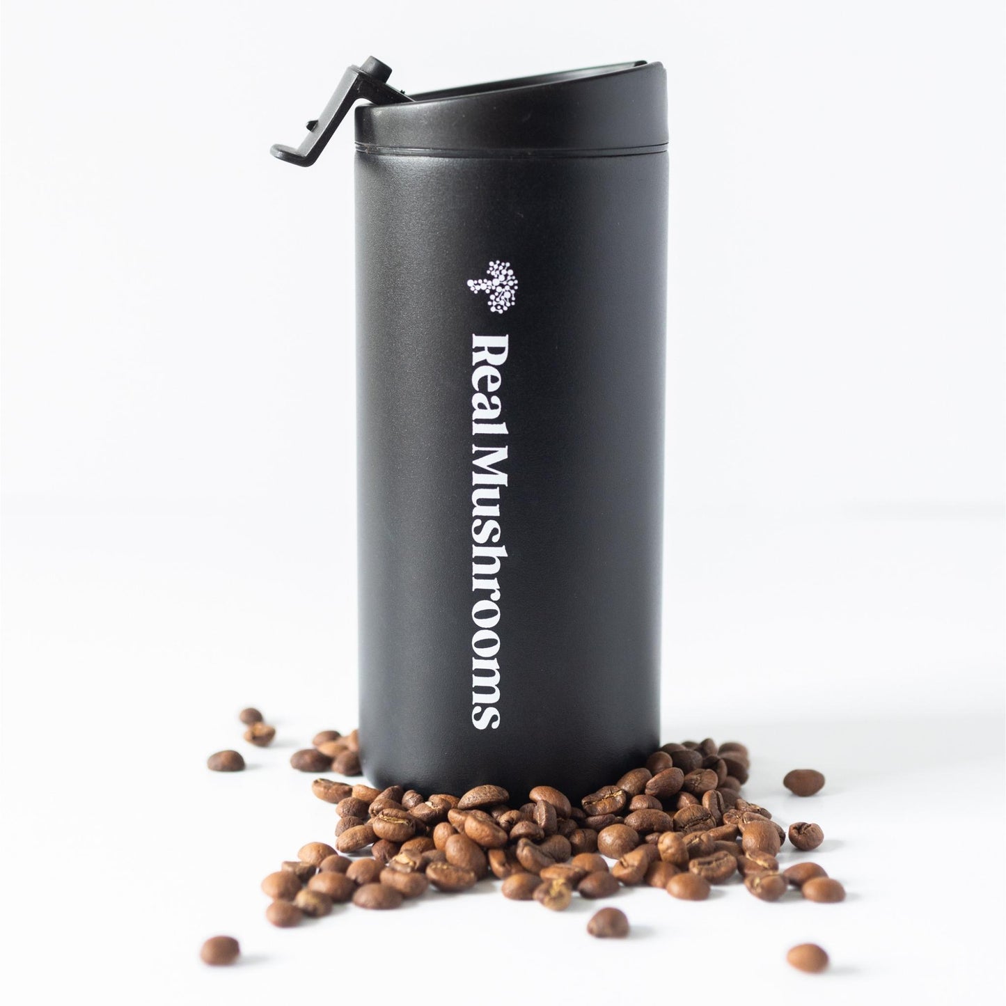 A black 12oz Travel Tumbler from Real Mushrooms stands upright with its lid open, surrounded by scattered coffee beans on a white background.