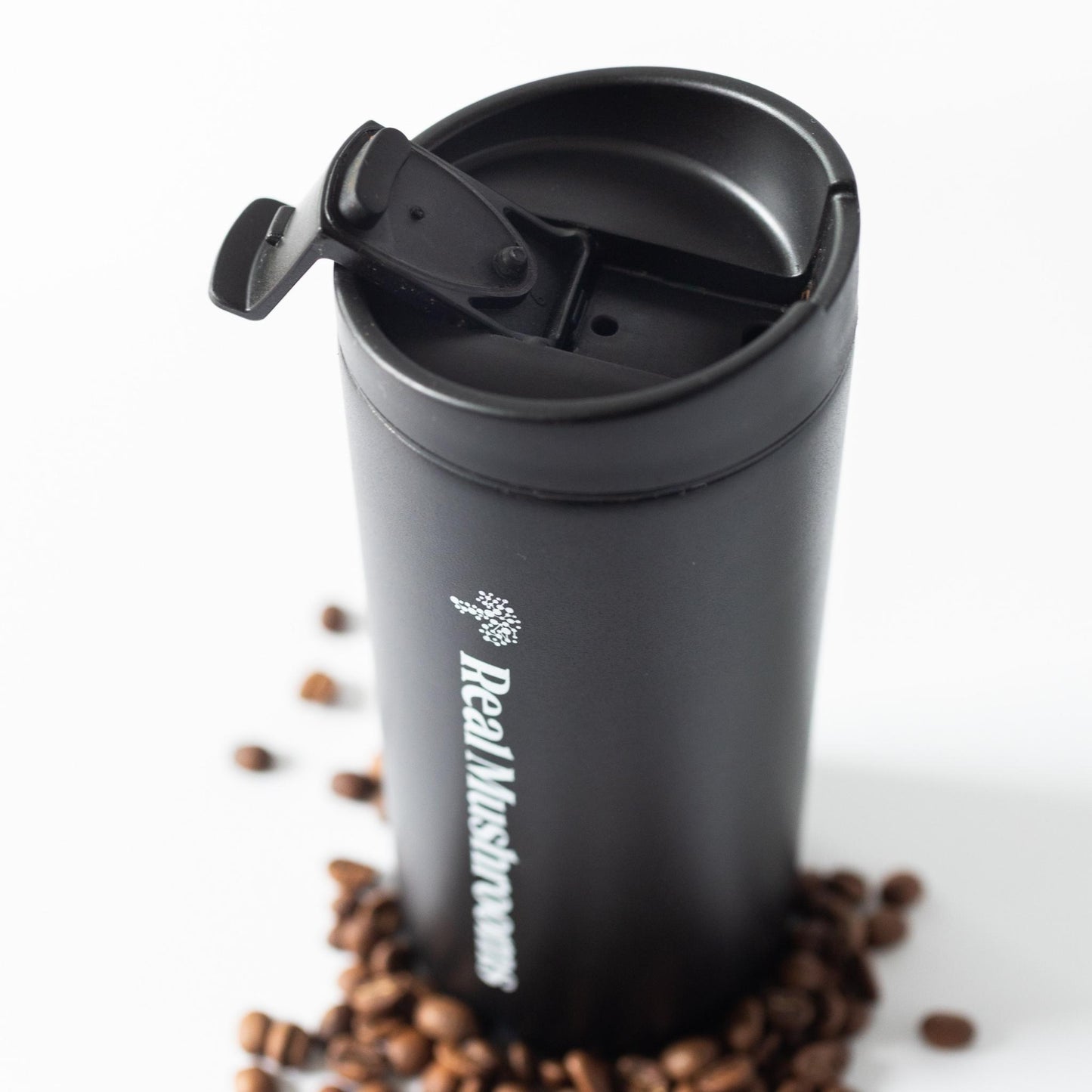 The Real Mushrooms 12oz Travel Tumbler, a black insulated, leak-proof mug with a flip-top lid, is shown on a white background surrounded by scattered coffee beans.