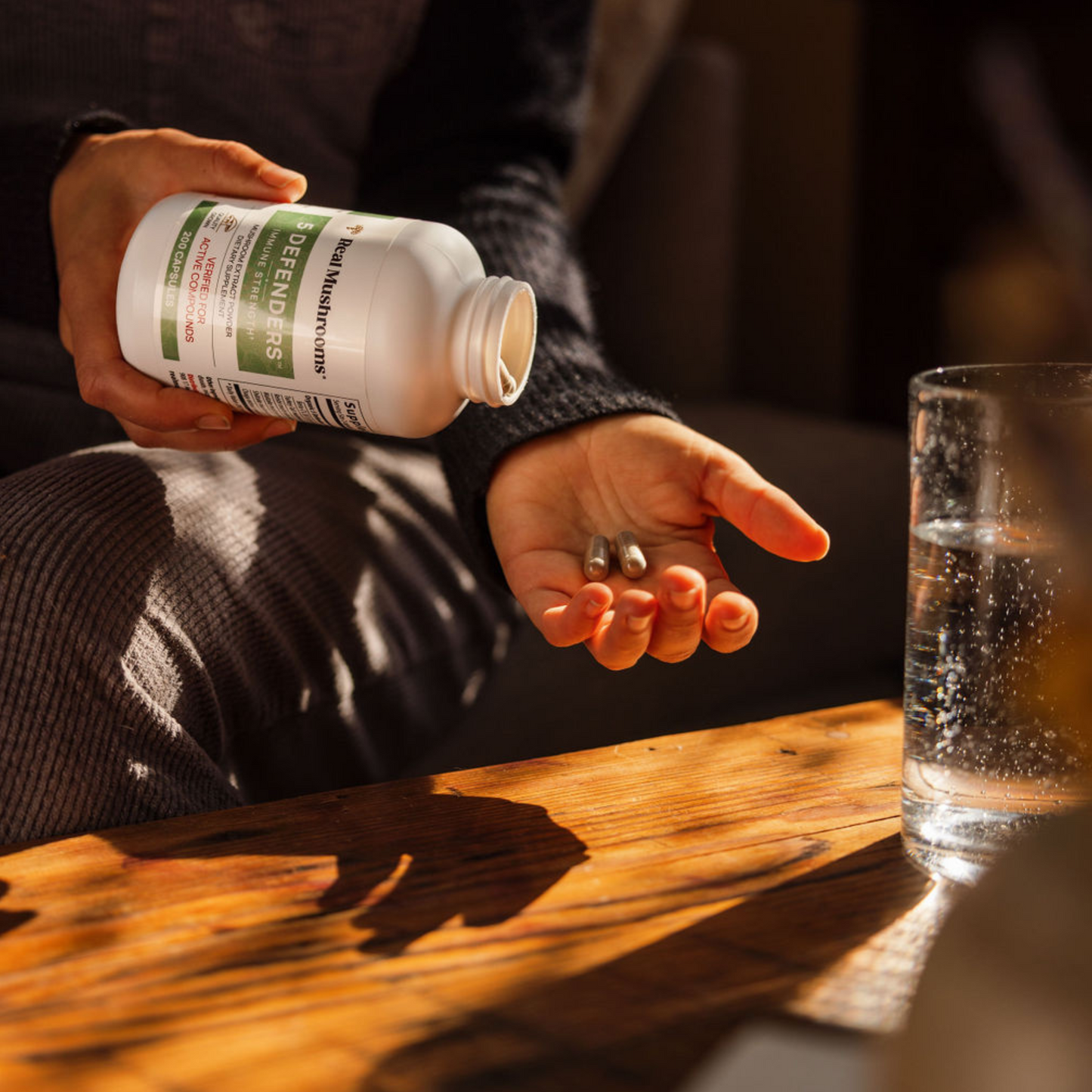 Someone pours 5 Defenders® Organic Mushroom Blend Capsules by Real Mushrooms from a white bottle into their hand, next to a glass of water on a wooden table.