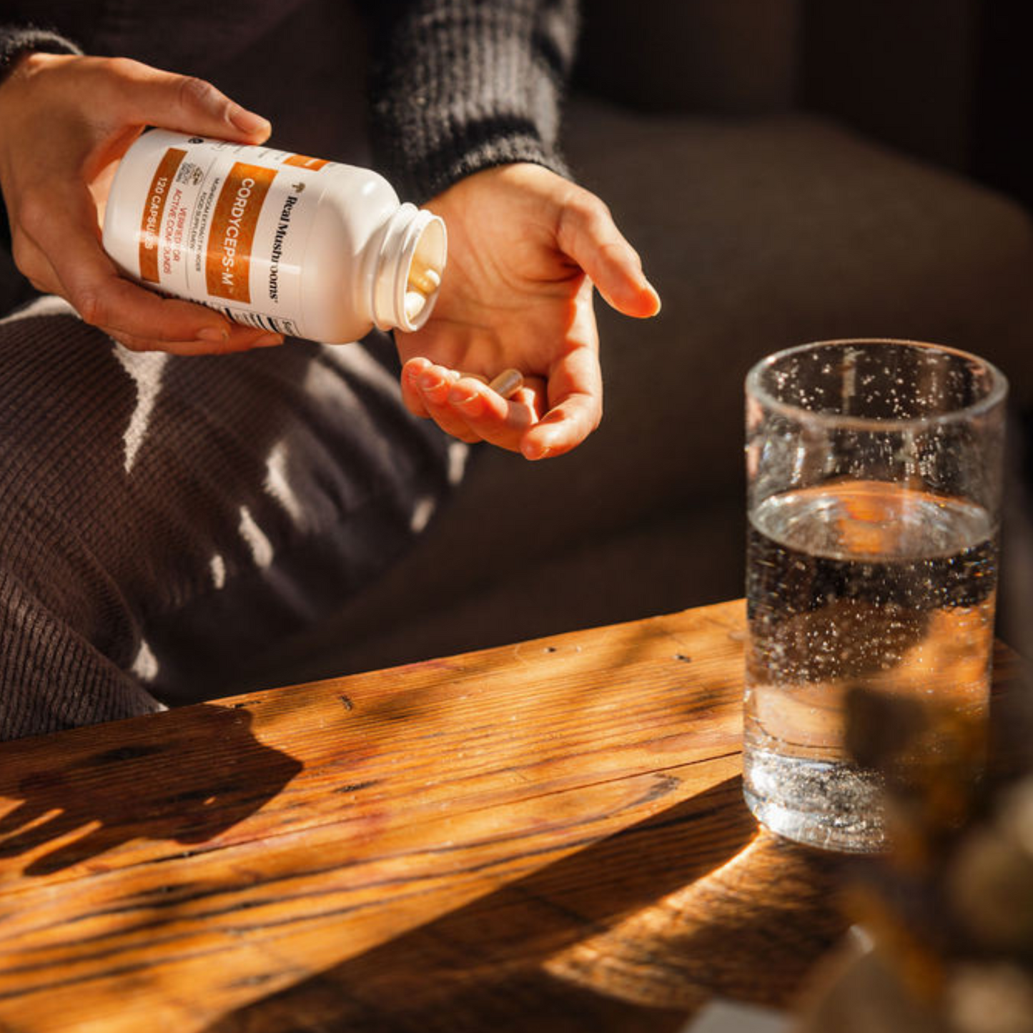 A person pours Real Mushrooms Organic Cordyceps Extract Capsules into their hand beside a glass of water on a wooden table, seeking natural energy and stamina support.