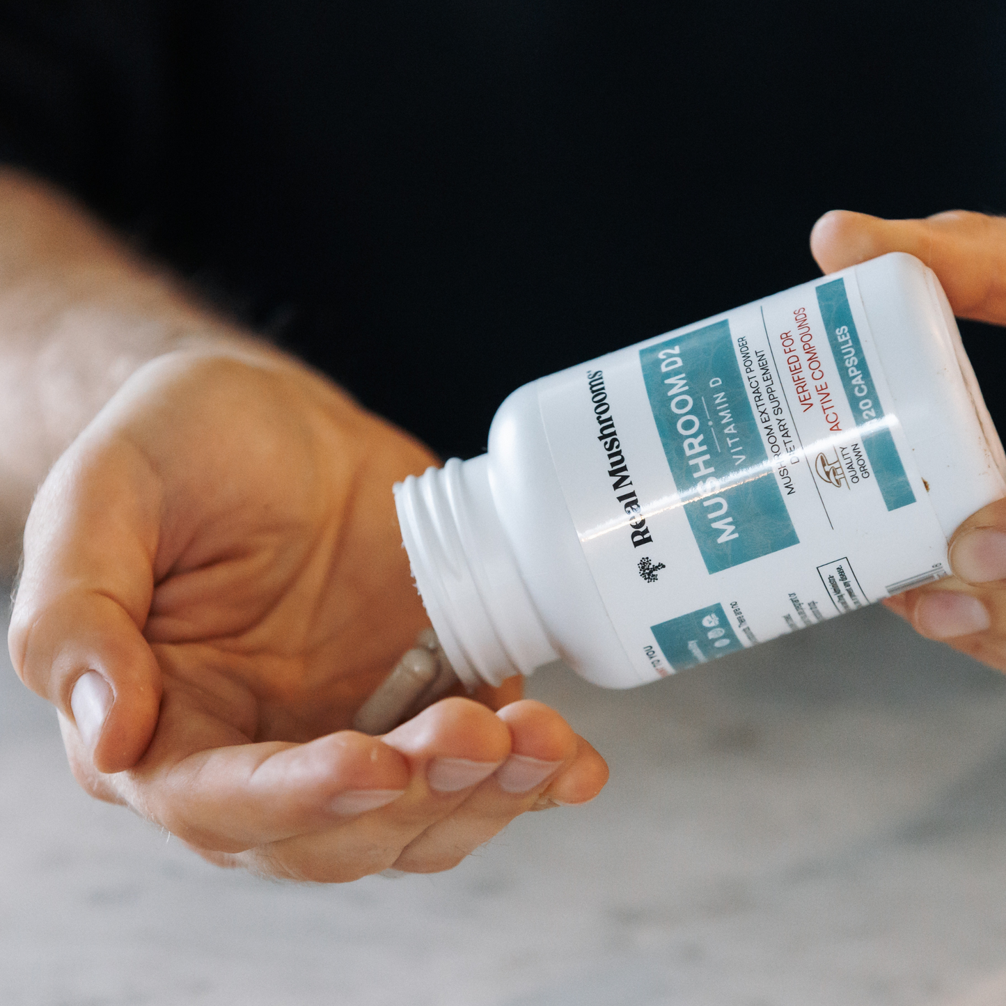 A person pours capsules from a white "Vitamin D from Organic Mushrooms" bottle labeled "Real Mushrooms" into their hand.
