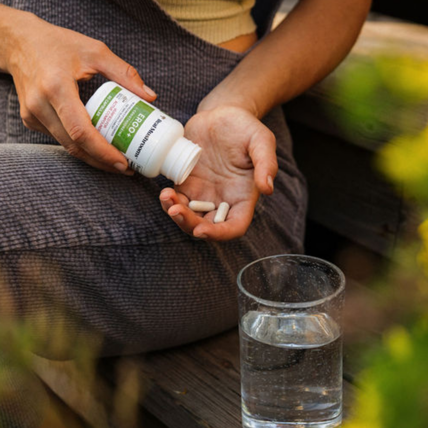 A person pours two white Ergo+ Ergothioneine Supplement tablets by Real Mushrooms from a bottle into their hand, with a glass of water on a wooden surface nearby.