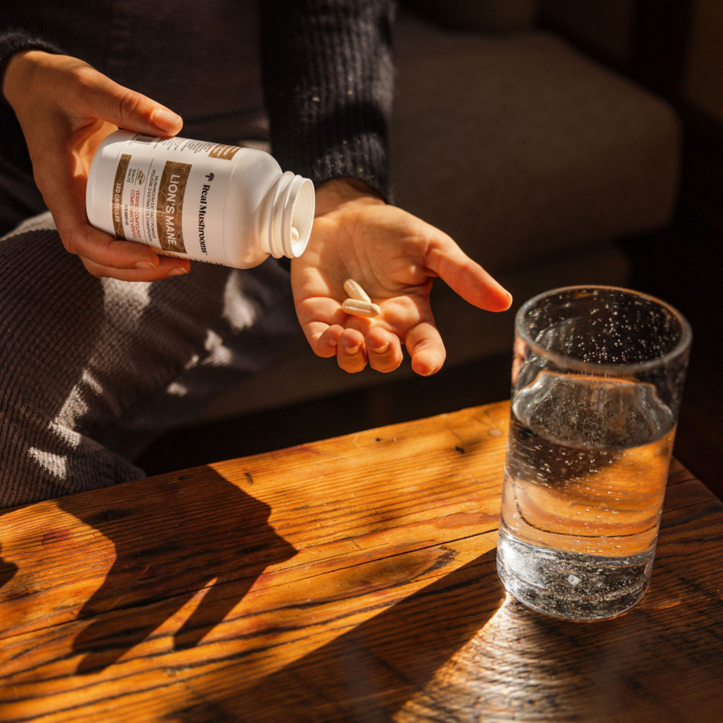 Someone pours Real Mushrooms Organic Lion's Mane Extract Capsules from the bottle into their hand beside a glass of water on a wooden table, supporting brain health.