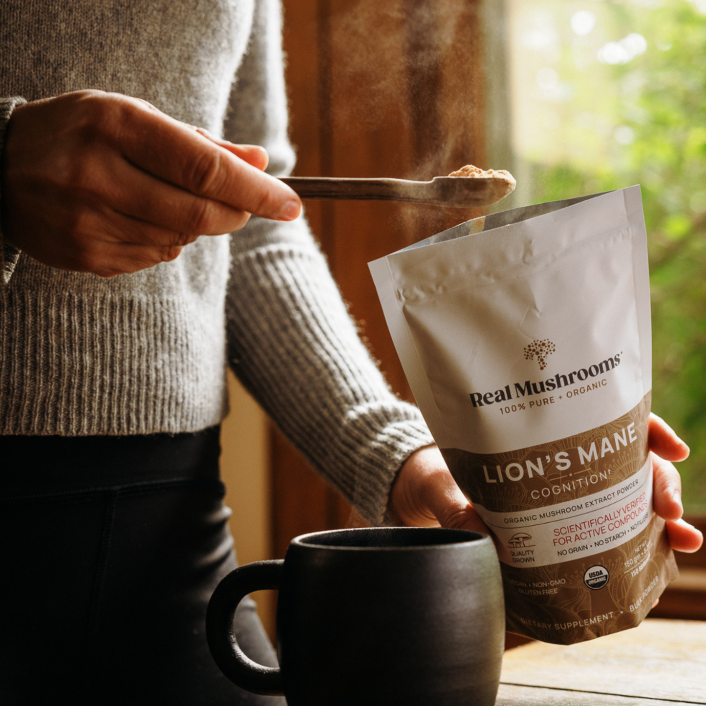 A person scoops Real Mushrooms Lion’s Mane Mushroom Powder into a black mug by a sunlit window, supporting immune health.