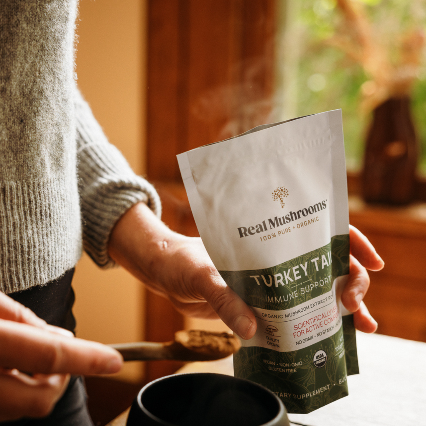 Someone scoops Real Mushrooms Turkey Tail Extract - Bulk Powder from a bag into a black mug in a warmly lit room.
