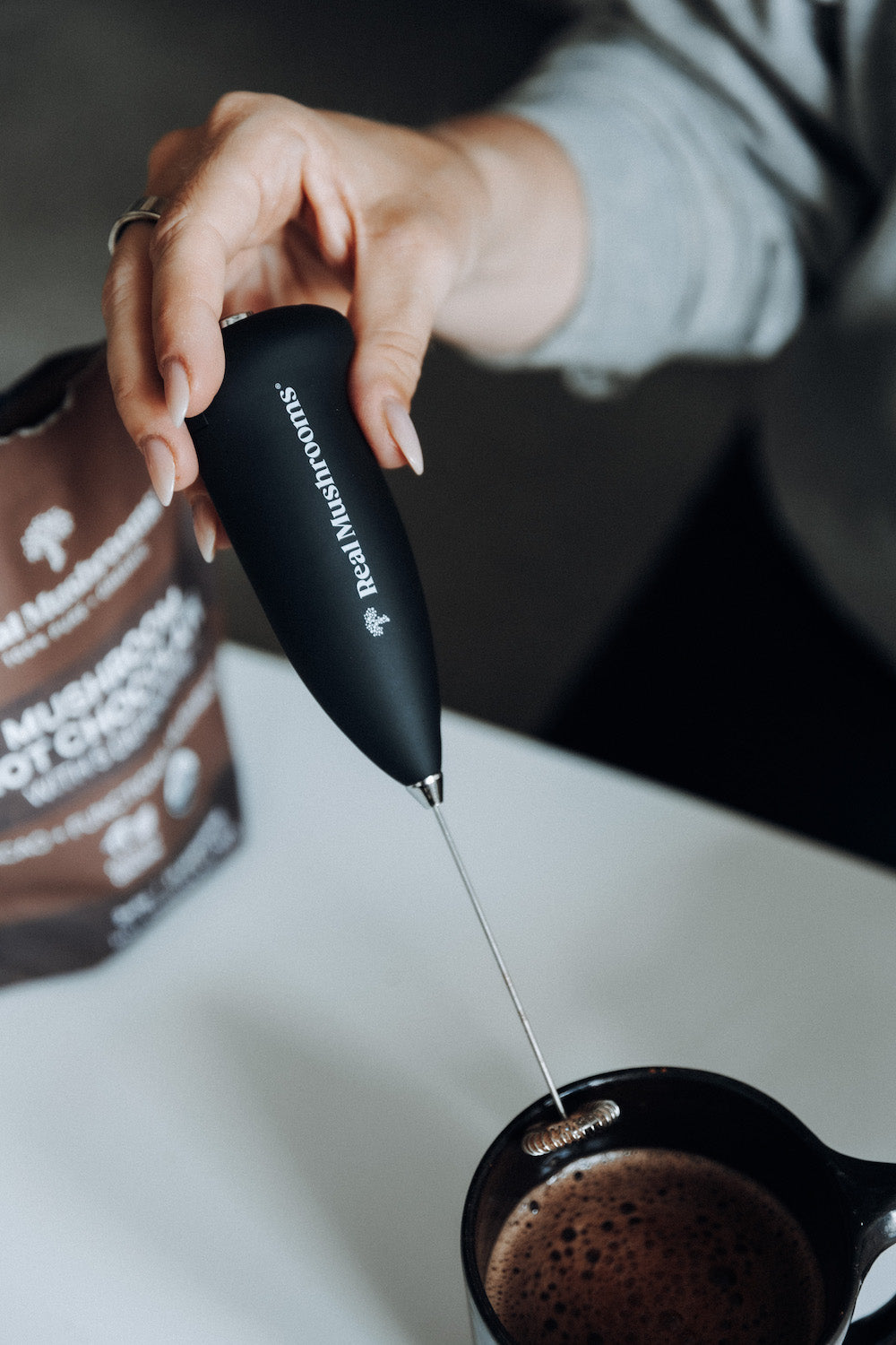 Someone uses the Real Mushrooms Electric Milk Frother with a stainless steel whisk to blend a drink in a black mug, while a "Mushroom" bag from Real Mushrooms sits on the table, hinting at tasty mushroom beverages.