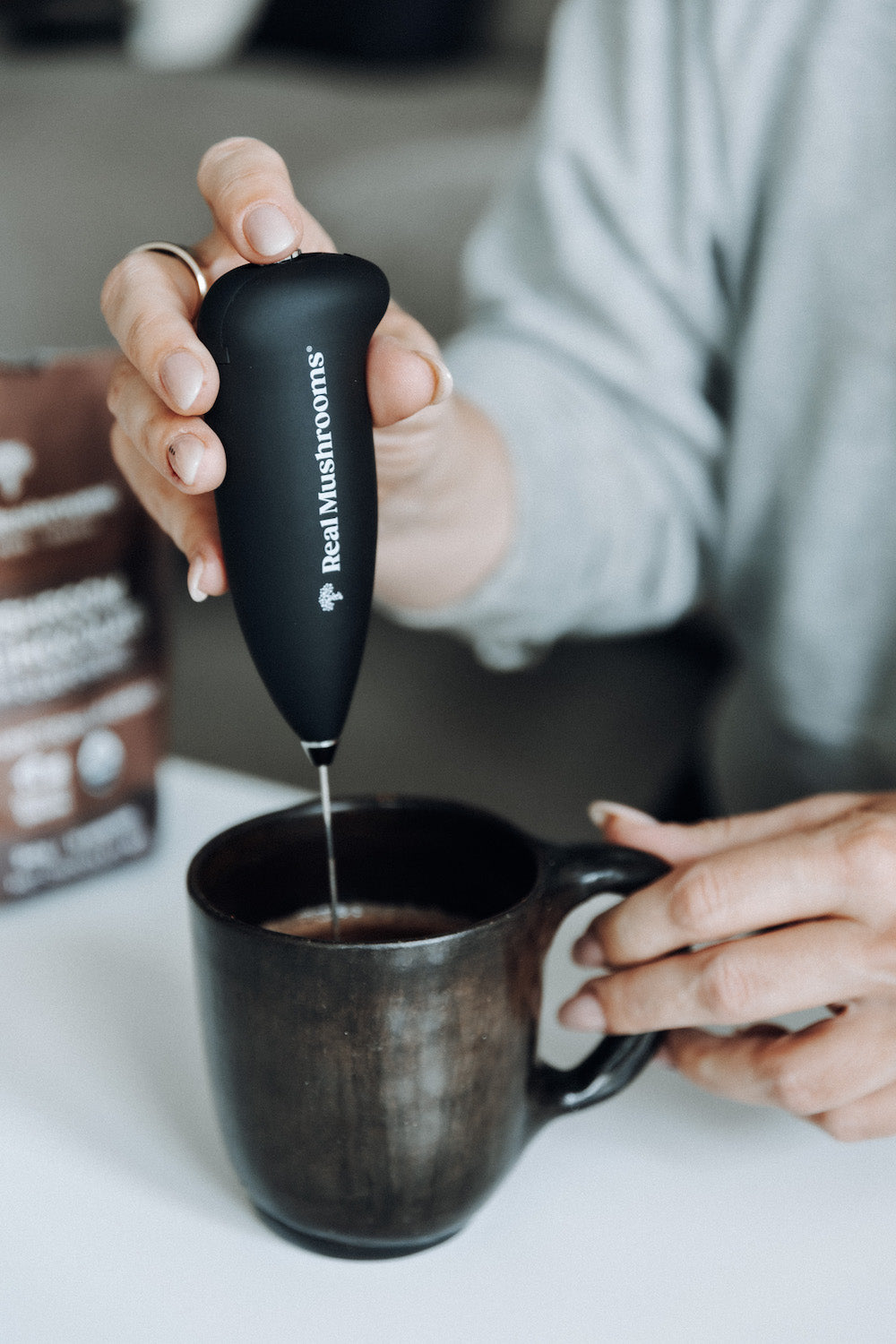 A person uses a Real Mushrooms Electric Milk Frother with a stainless steel whisk to blend a drink in a dark ceramic mug, ideal for making mushroom beverages.