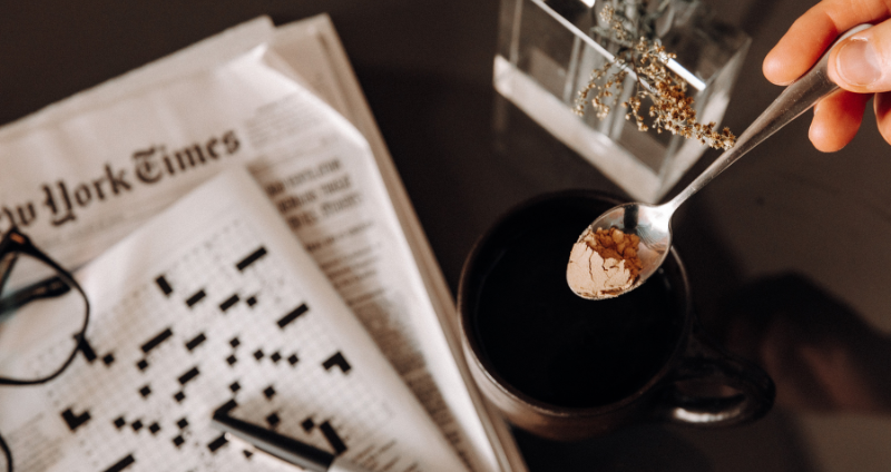 A hand holds a spoon over a dark mug with a newspaper crossword, glasses, and pen on a table. Nearby, an open bottle of the best supplements for focus and concentration hints at an early start to a productive day.
