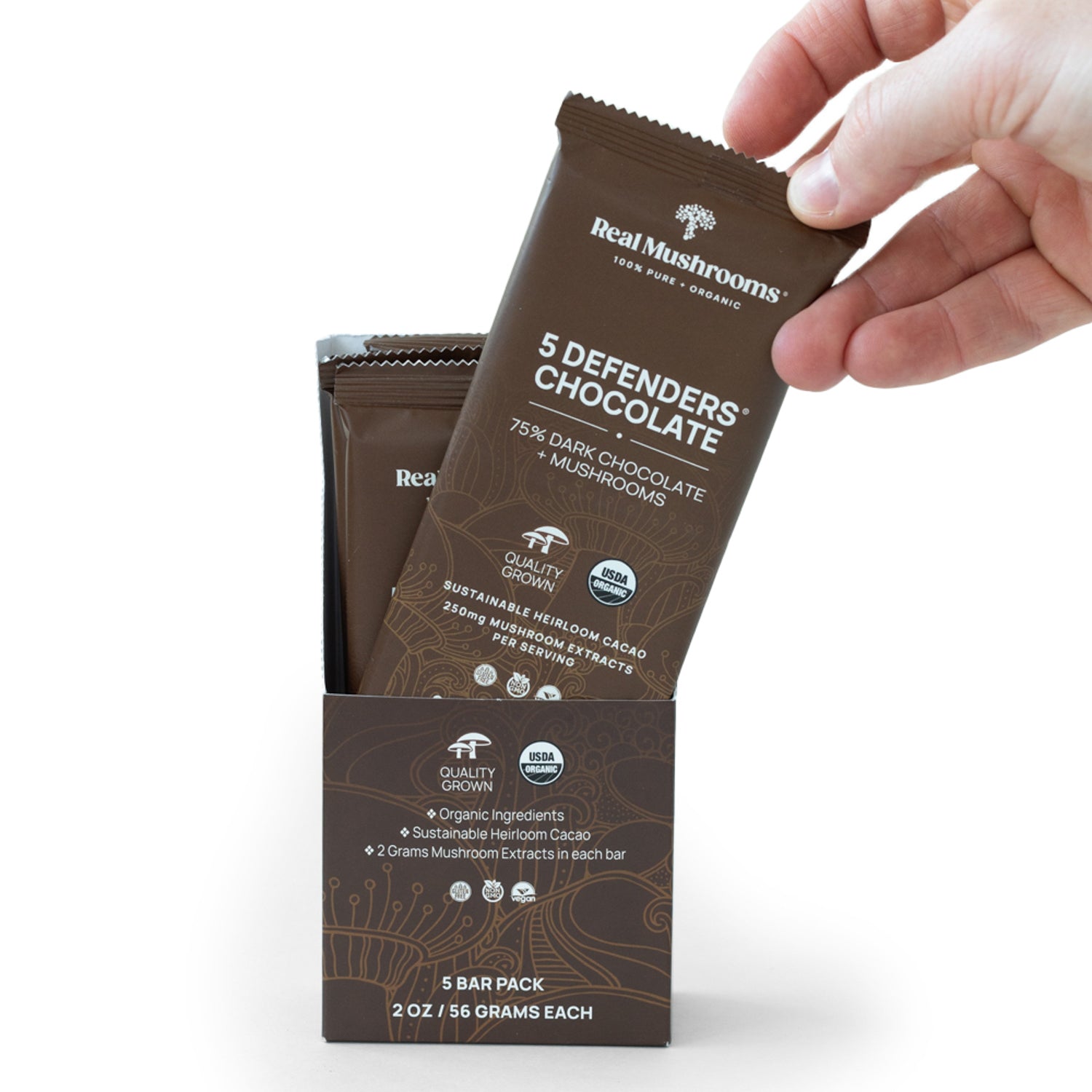 A hand holds a chocolate bar labeled "Real Mushrooms - Dark Chocolate" infused with organic mushroom extracts. Each vegan-friendly bar is packaged in a brown box, proudly highlighting its commitment to sustainable ingredients, coming as part of the Mushroom Chocolate Bar - 5 Pack.