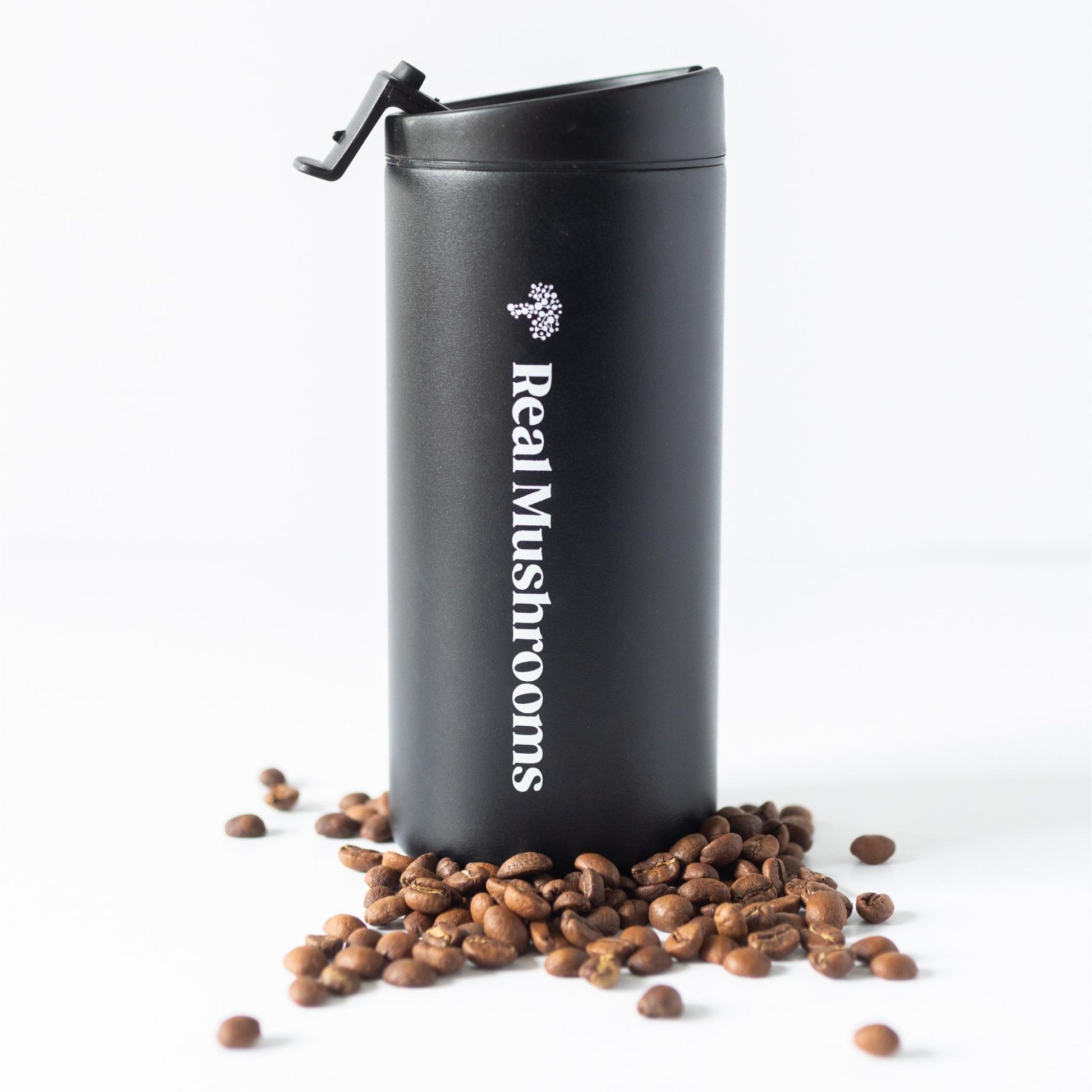 A black 12oz Travel Tumbler from Real Mushrooms stands upright with its lid open, surrounded by scattered coffee beans on a white background.