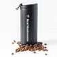 A black 12oz Travel Tumbler from Real Mushrooms stands upright with its lid open, surrounded by scattered coffee beans on a white background.