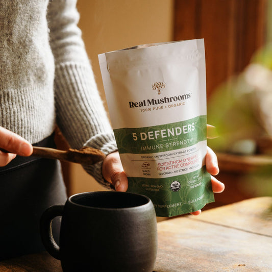 Someone scoops 5 Defenders® Organic Mushroom Complex – Bulk Powder by Real Mushrooms from a pouch into a black mug on a wooden table.