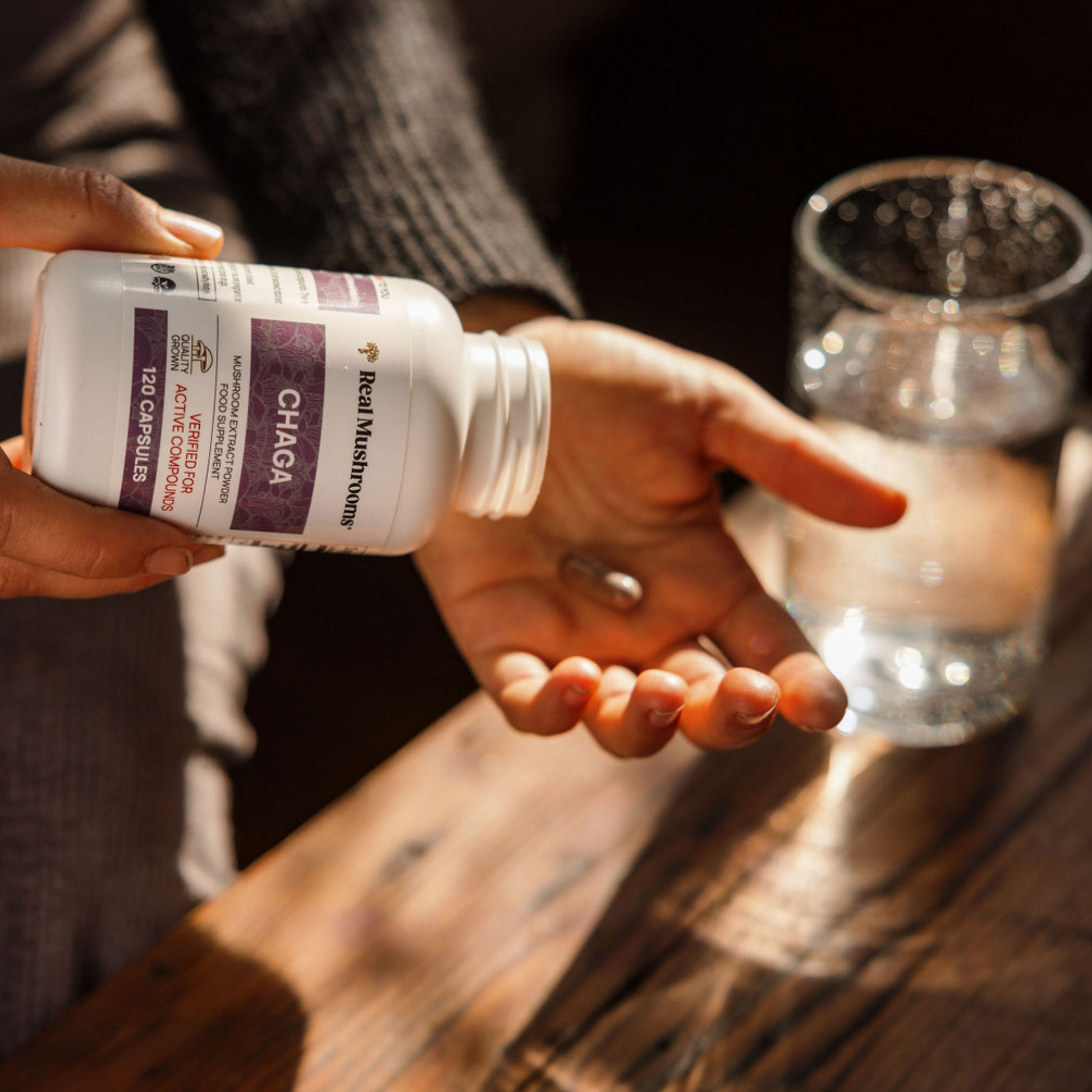A person pours a Real Mushrooms Organic Chaga Extract Capsule from a bottle into their hand, with a glass of water on a wooden table nearby, supporting immune health.