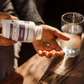 A person pours a Real Mushrooms Organic Chaga Extract Capsule from a bottle into their hand, with a glass of water on a wooden table nearby, supporting immune health.