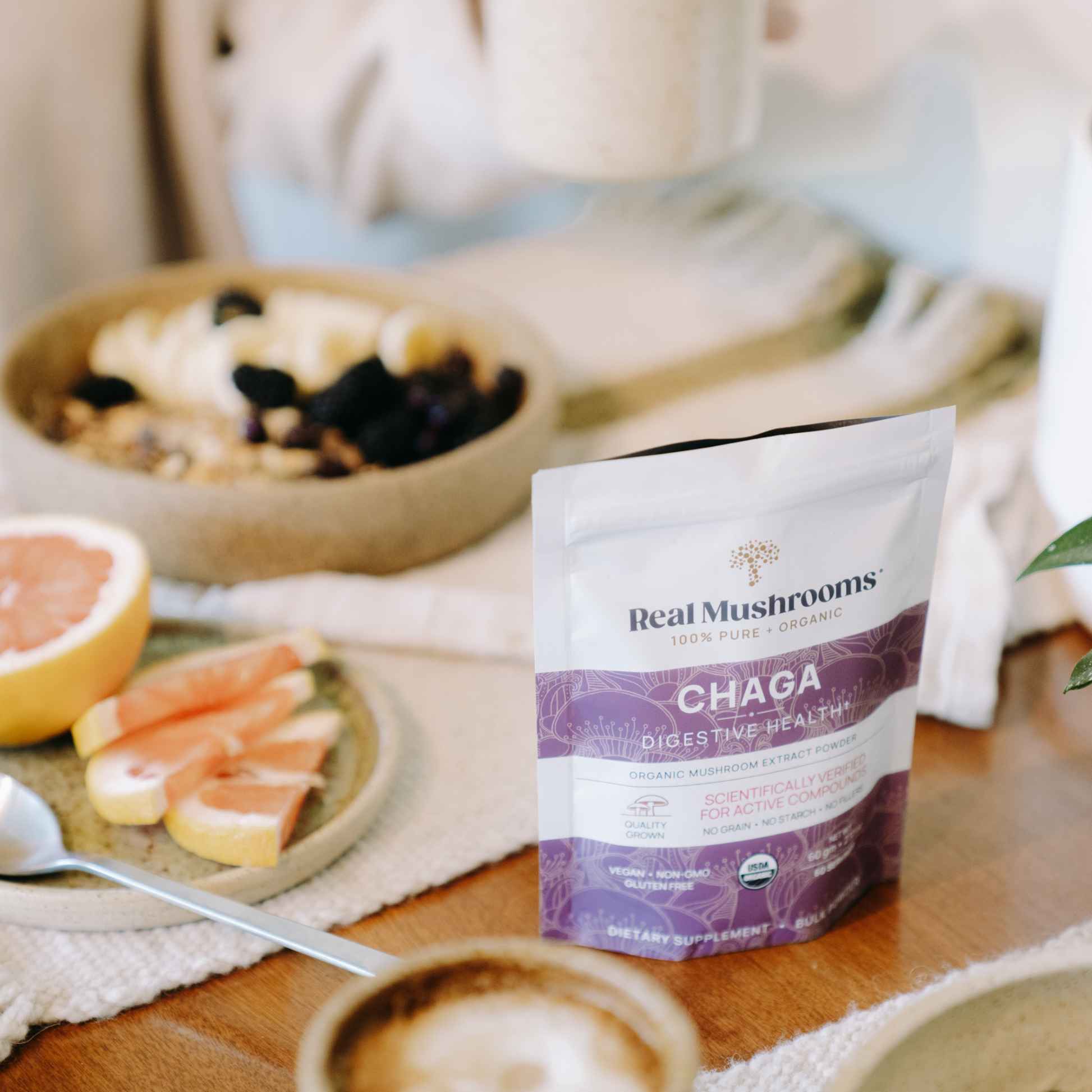 A packet of Real Mushrooms Organic Chaga Extract Powder sits on a table beside fruit, sliced grapefruit, and coffee—ideal for daily immune support.