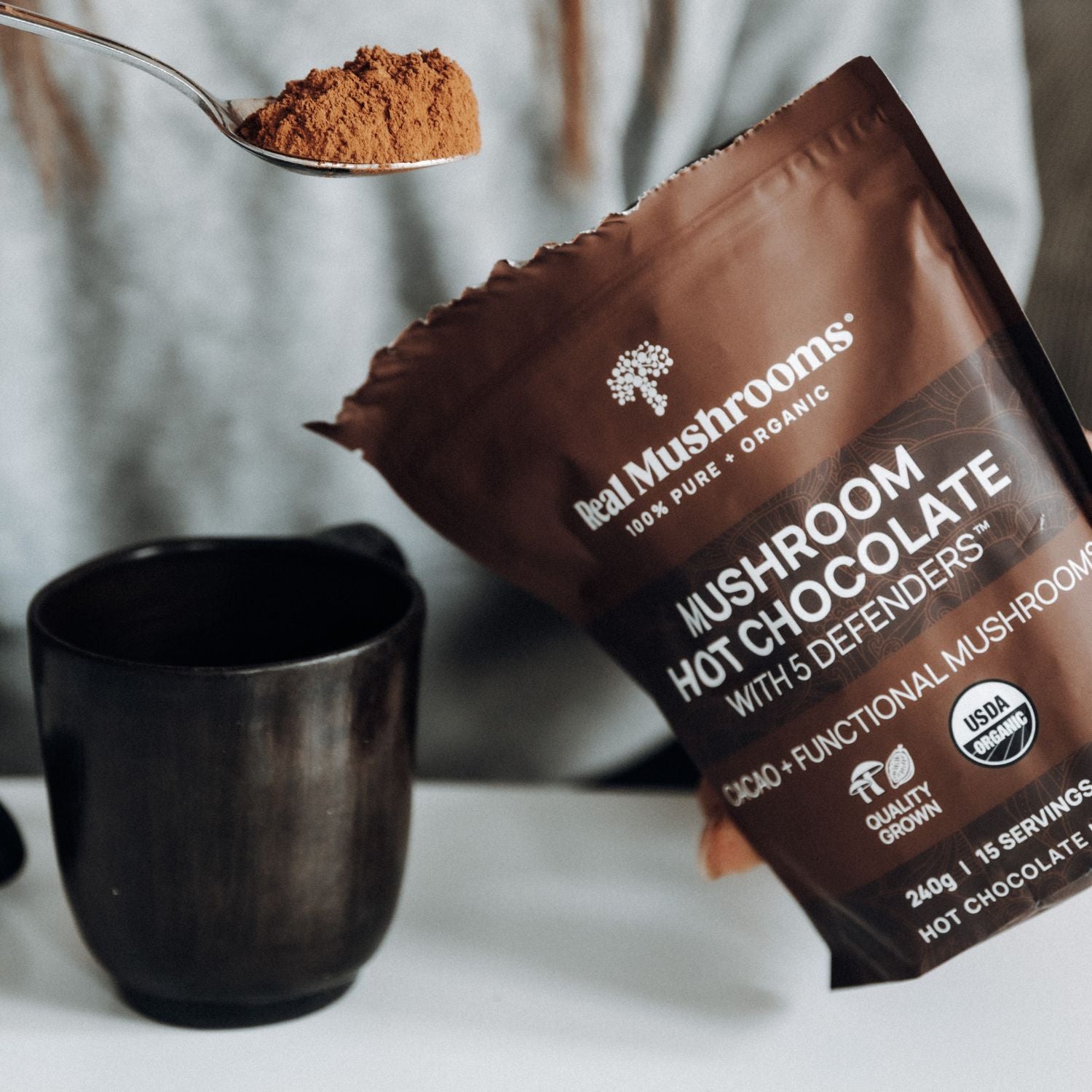 A person holds a packet of Real Mushrooms Mushroom Hot Chocolate Mix, a vegan blend made with organic mushroom extracts, while scooping the powder into a black mug.
