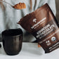 A person holds a packet of Real Mushrooms Mushroom Hot Chocolate Mix, a vegan blend made with organic mushroom extracts, while scooping the powder into a black mug.