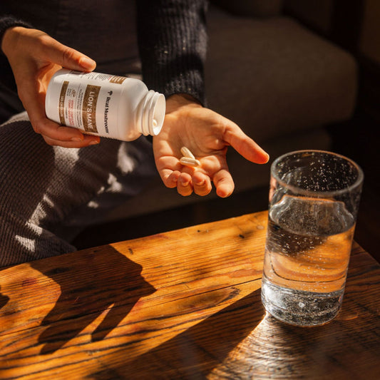A person pours Real Mushrooms Organic Lion's Mane Extract Capsules from a bottle into their hand beside a glass of water on a wooden table, showcasing immune support benefits from organic mushroom extracts.