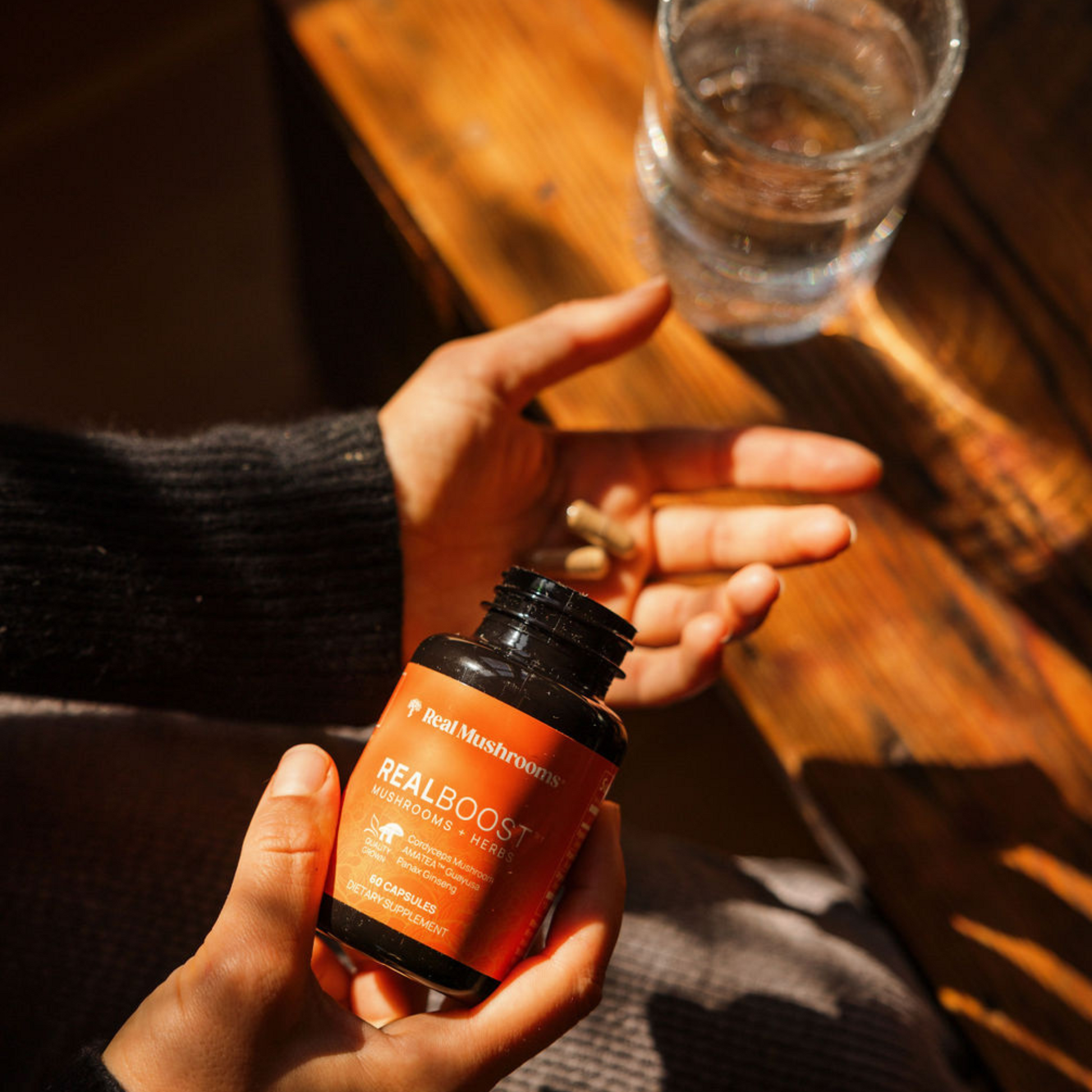 A person holds a Real Mushrooms RealBoost bottle—containing cordyceps, guayusa, and ginseng—and two capsules in one hand, with a glass of water on a wooden surface nearby.