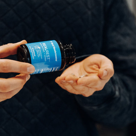 A person pours capsules from a Real Mushrooms RealRest bottle—supplemented with Reishi, Valerian, and Lemon Balm—into their hand.