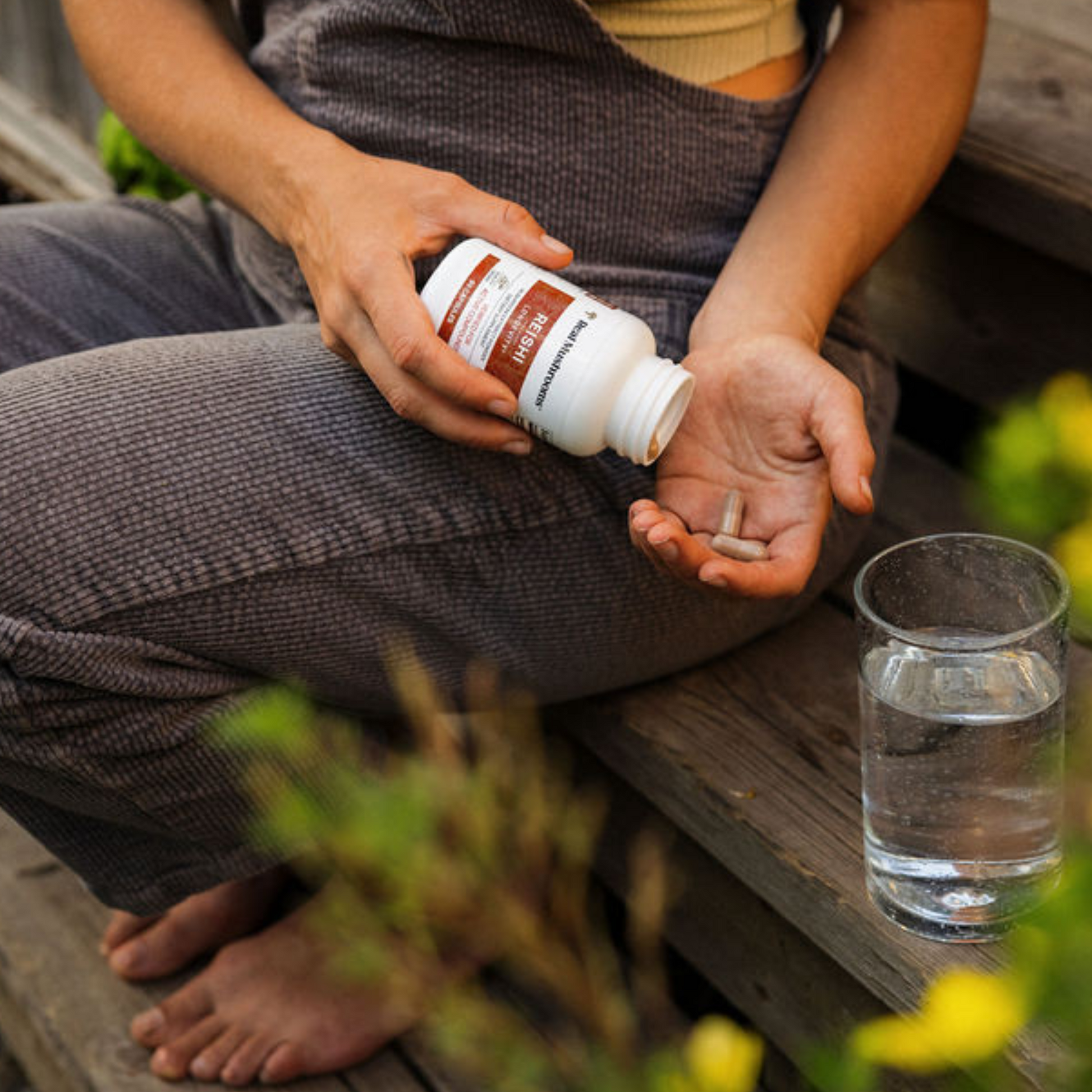 A person on outdoor wooden steps pours Real Mushrooms Organic Reishi Mushroom Capsules into their hand from a supplement bottle, with a glass of water nearby.