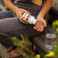 A person sits on wooden steps pouring Real Mushrooms Organic Reishi Mushroom Capsules from the bottle into their hand, with a glass of water nearby.