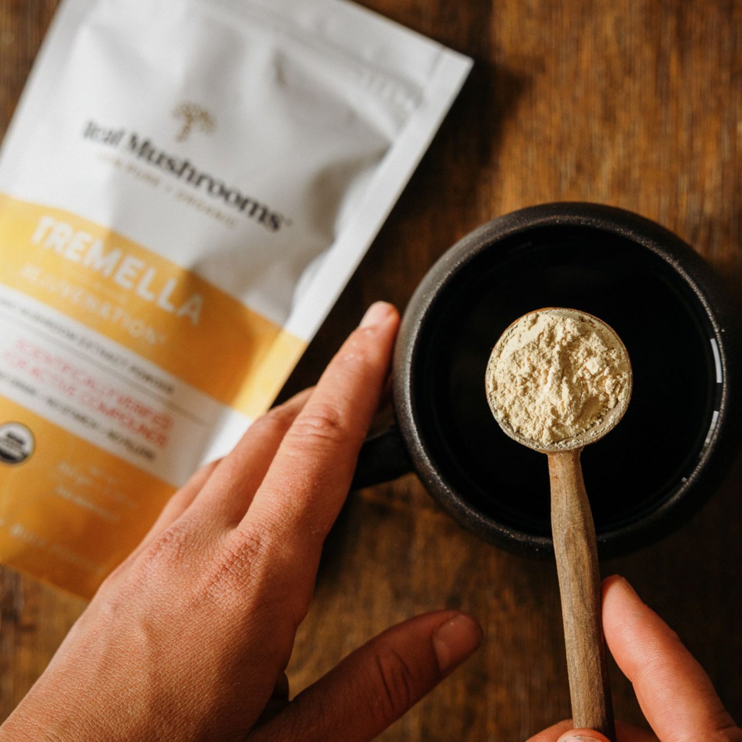 A hand holds a wooden spoon with Organic Tremella Extract Powder above a dark mug, with a Real Mushrooms pouch in the background on a wooden surface, showcasing natural immune support.
