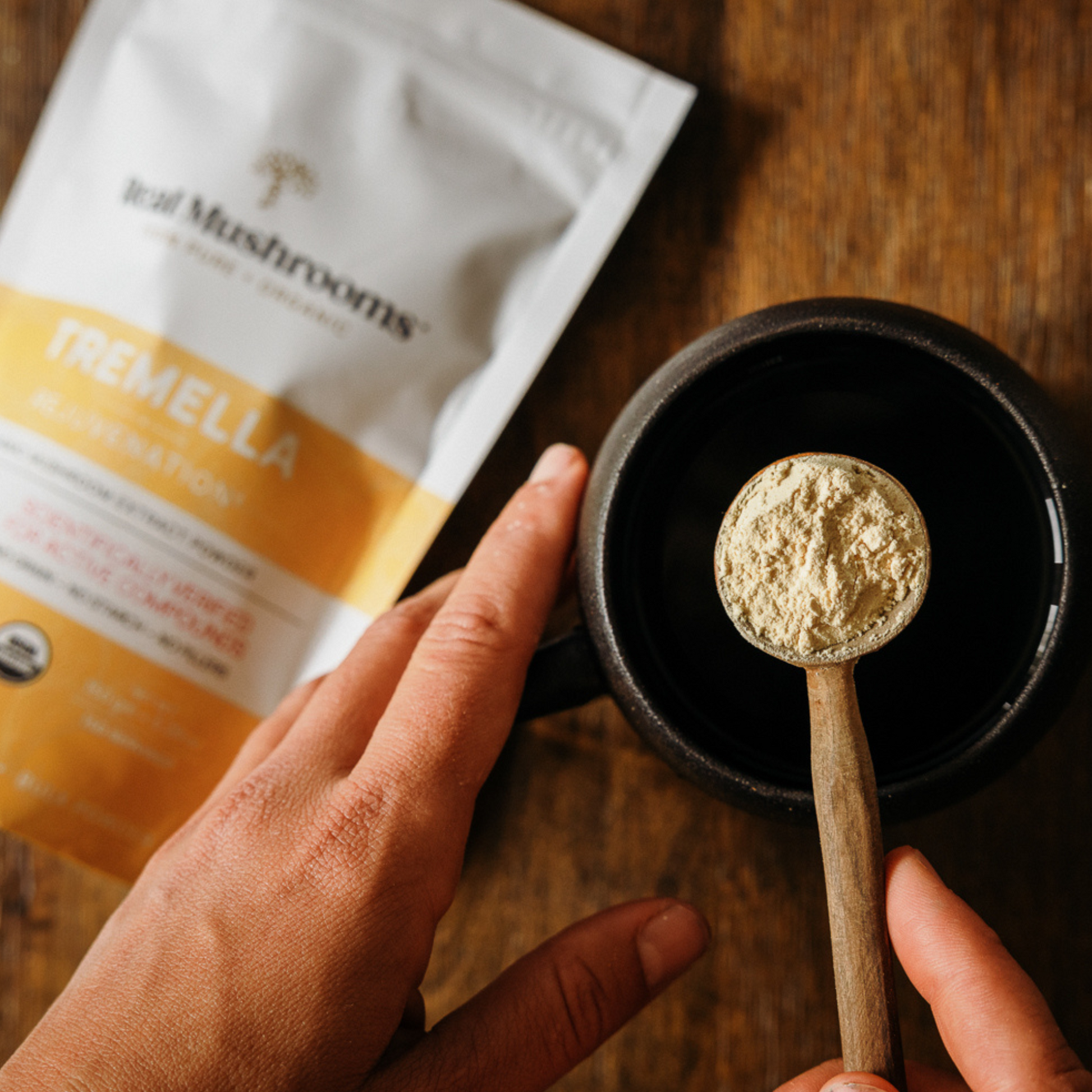 A hand holds Real Mushrooms Organic Tremella Extract Powder above a black mug, with the product packet in the background—perfect for nourishing skin, hair, and nails.