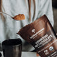 A person holds a spoonful of USDA Certified Organic Hot Chocolate Sample powder above a black mug, with a brown packet labeled "Real Mushrooms" nearby.