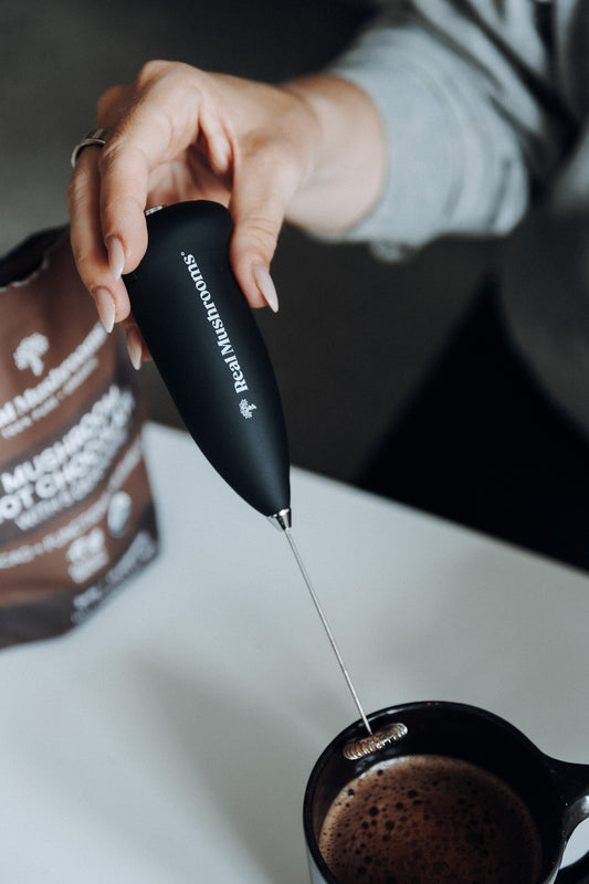 Someone uses the Real Mushrooms Electric Milk Frother with a stainless steel whisk to blend a drink in a black mug, while a "Mushroom" bag from Real Mushrooms sits on the table, hinting at tasty mushroom beverages.