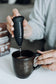 A person uses a Real Mushrooms Electric Milk Frother with a stainless steel whisk to blend a drink in a dark ceramic mug, ideal for making mushroom beverages.
