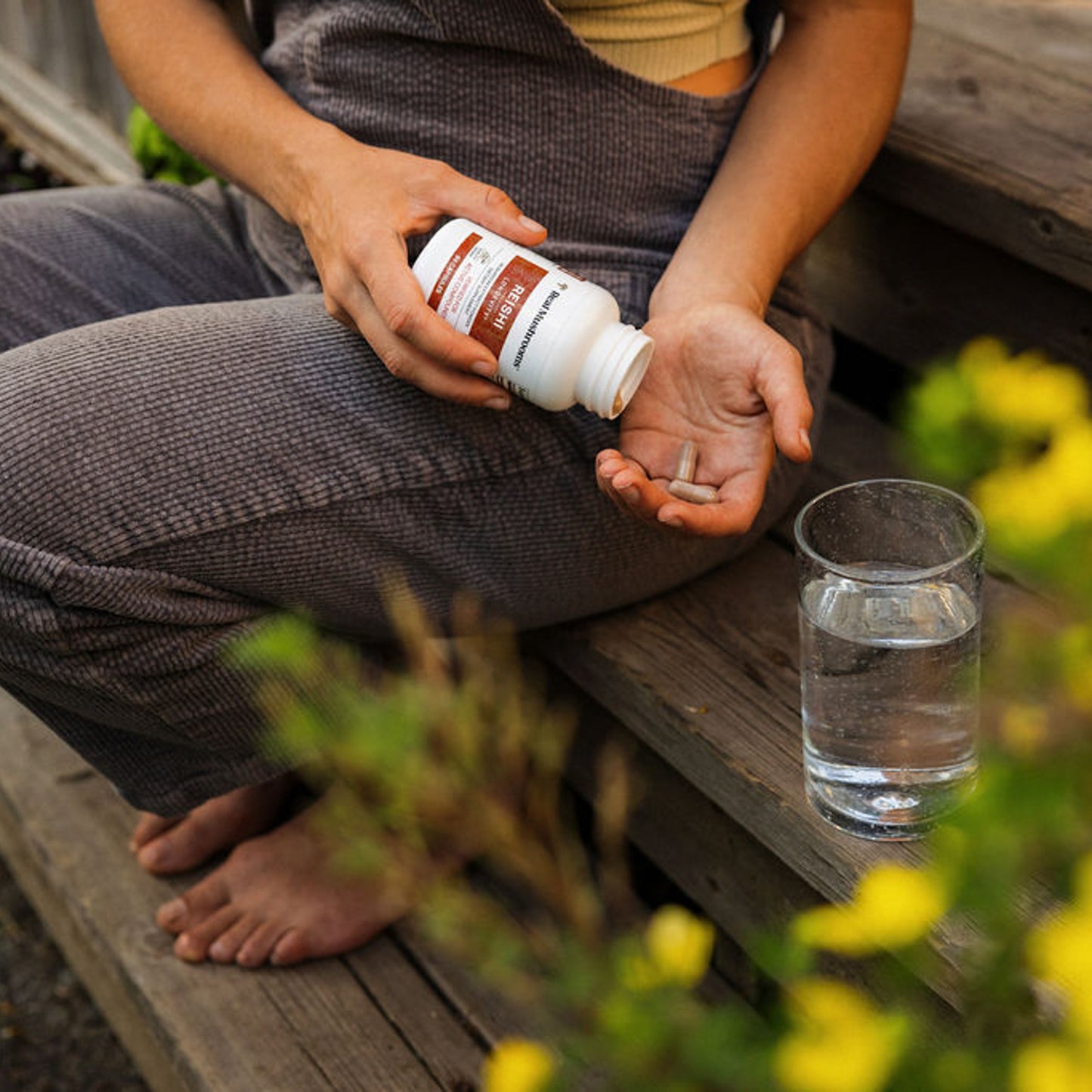 Sitting on wooden steps, a person holds a bottle of Real Mushrooms Organic Reishi Mushroom Capsules and a pill, while a glass of water sits beside them.