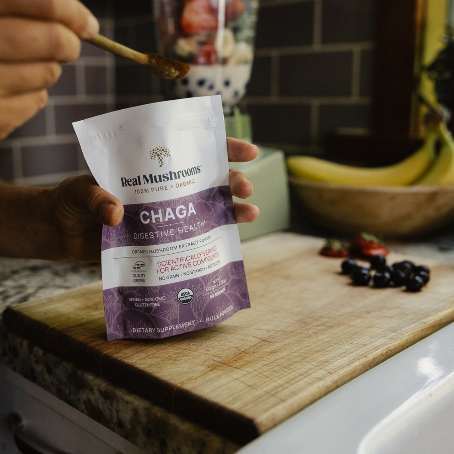 A person holds a bag of Real Mushrooms Organic Chaga Extract Powder on a kitchen counter. In the background, a blender filled with fruit awaits the addition of beta-glucans-rich goodness.