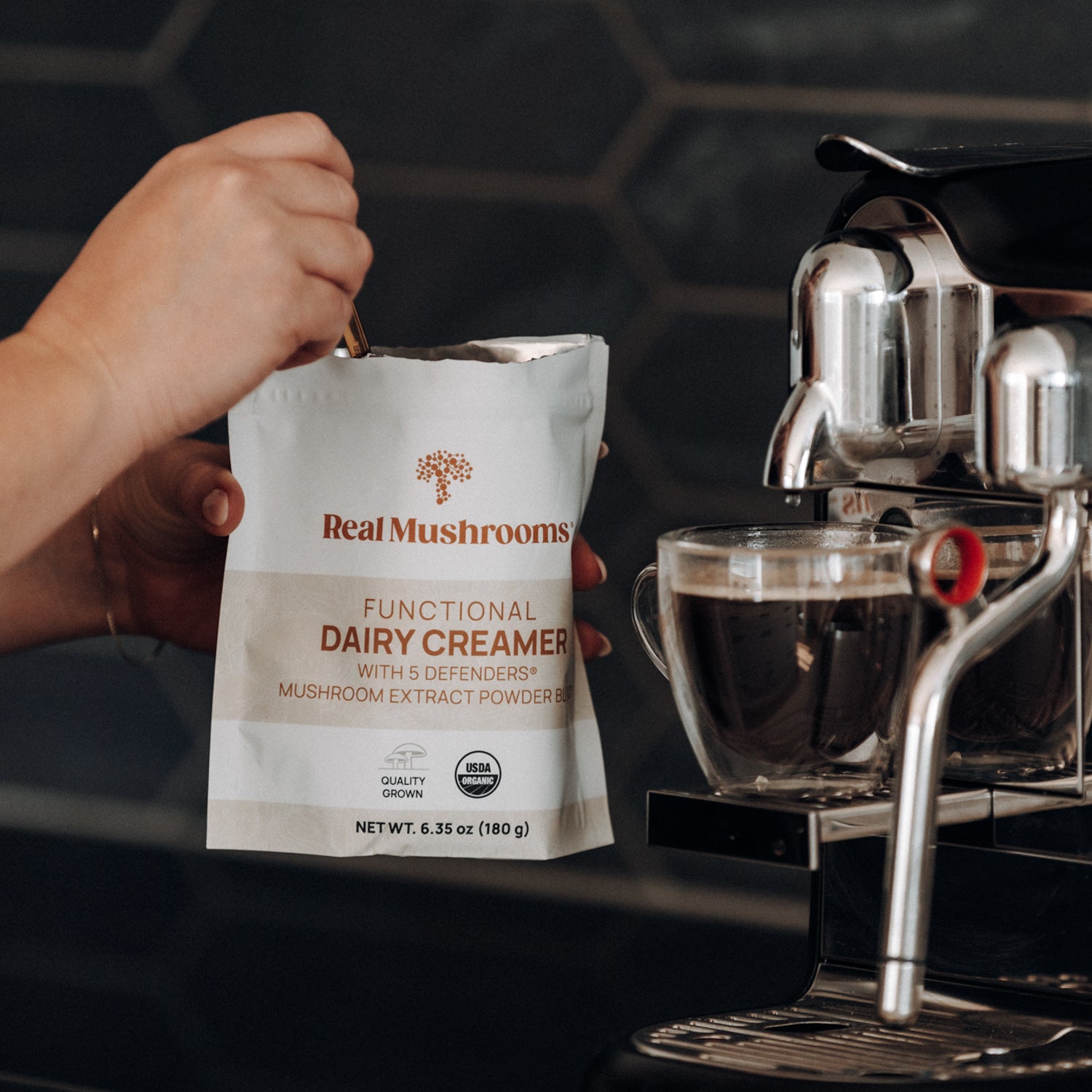 A hand scoops organic mushroom extract powder from a gluten-free "Functional Dairy Creamer Powder" bag by Real Mushrooms beside an espresso machine, with a glass cup waiting on the drip tray.