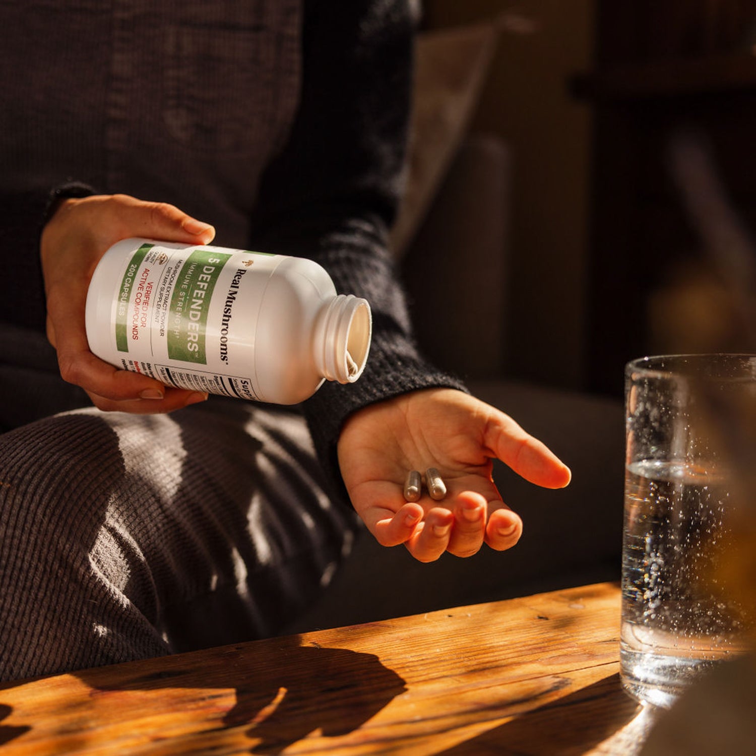 A person is holding a bottle of Real Mushrooms 5 Defenders Organic Mushroom Blend Capsules, pouring the gluten-free capsules into their other hand beside a glass of water on a wooden table.