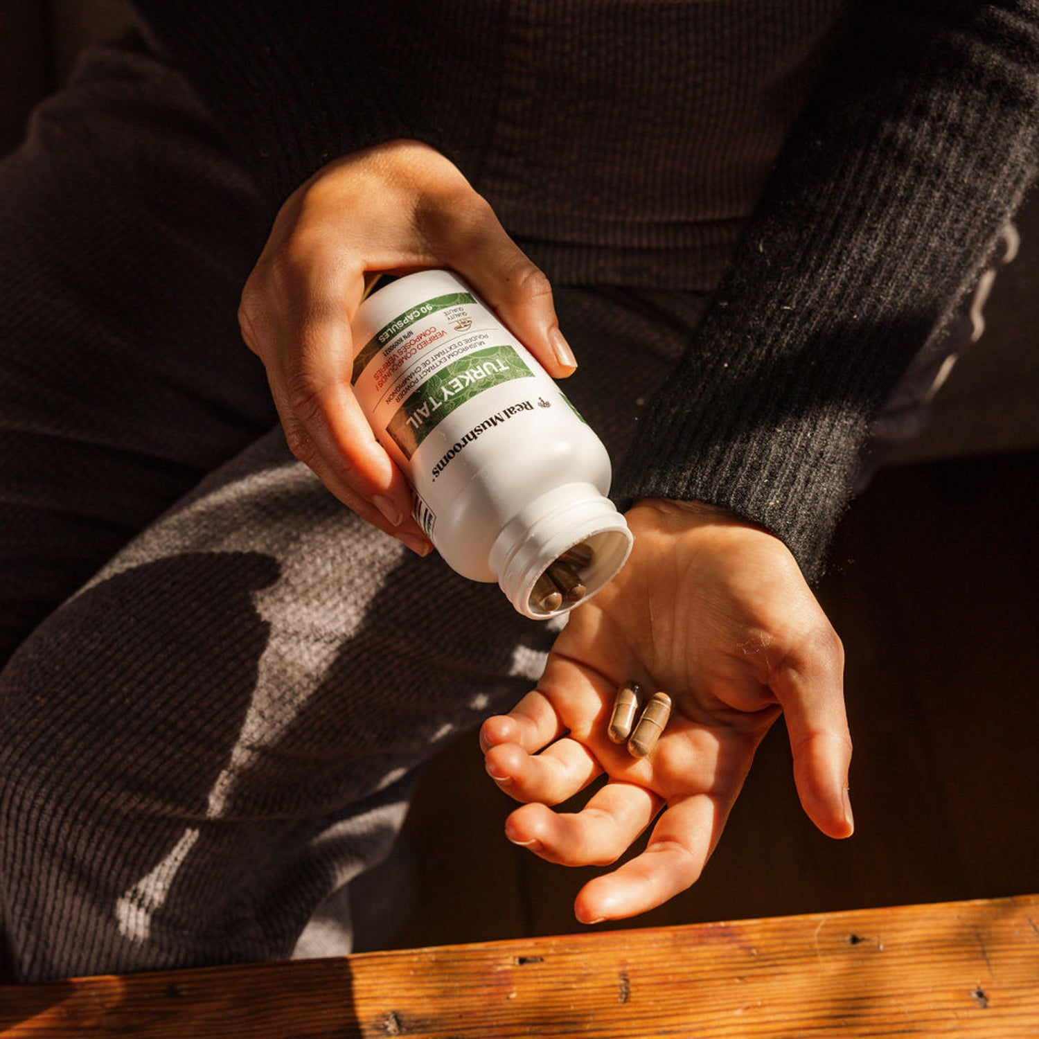 A person sits on a wooden surface, pouring Real Mushrooms' Turkey Tail Mushroom Capsules into their hand. Known for their high Beta-glucans content, these supplements provide a natural boost to wellness.