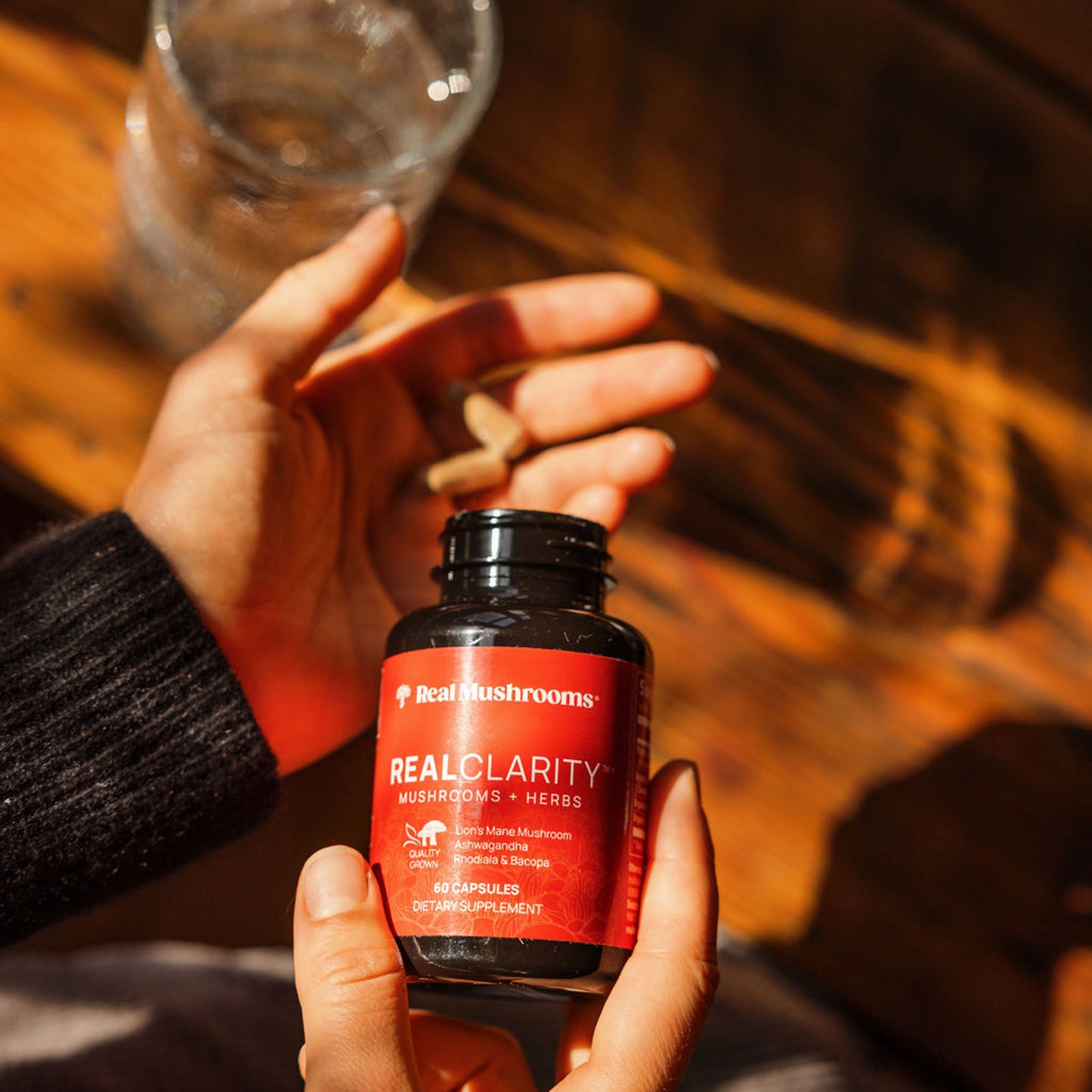 A person holds a bottle of "RealClarity" mushroom supplement capsules by Real Mushrooms, enriched with Lion's Mane, Ashwagandha, Rhodiola, and Bacopa, alongside a glass of water resting on the wooden surface.