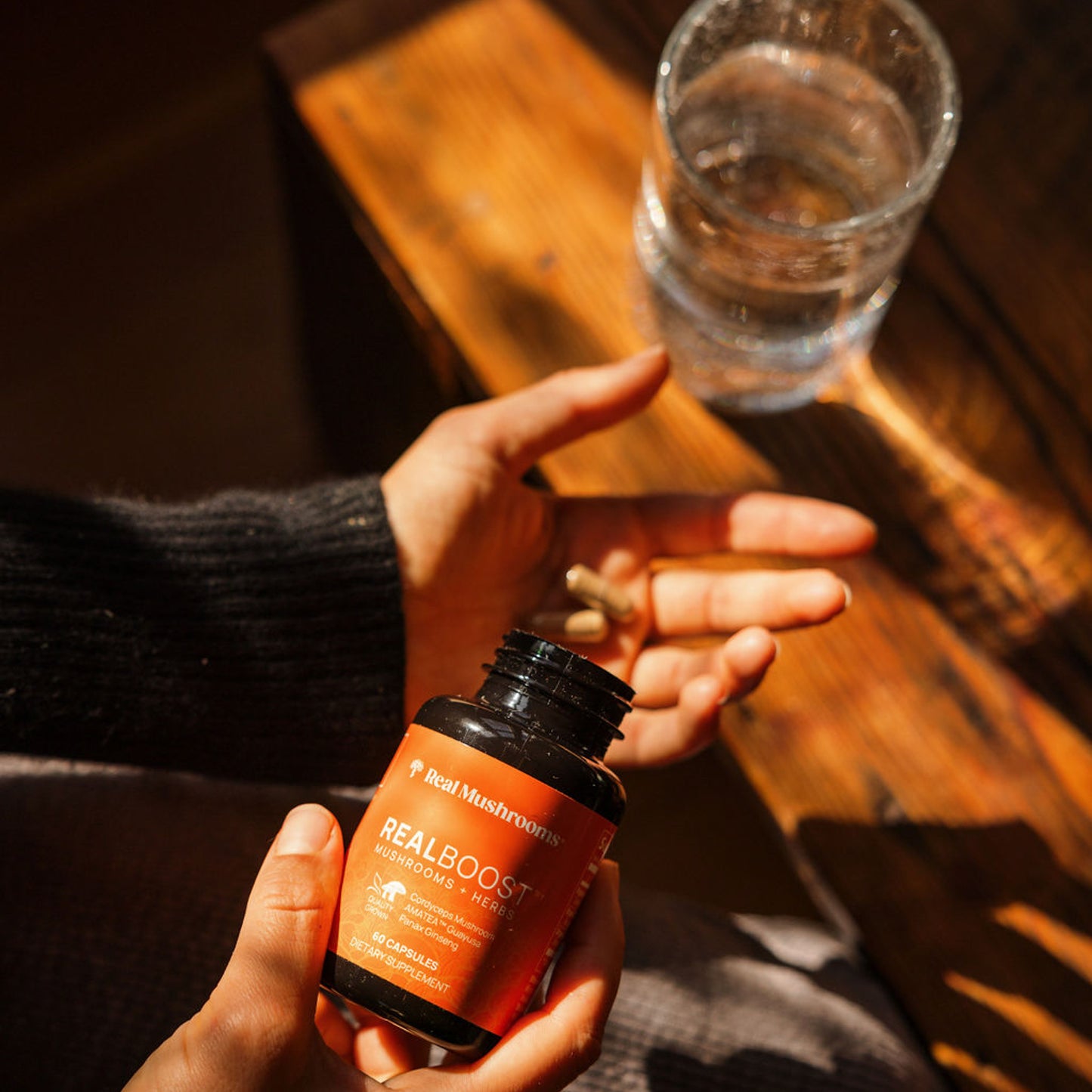 A person holds a bottle of Real Mushrooms Reishi capsules in one hand and two capsules in the other, with a glass of water resting on a sunlit wooden surface, alongside RealBoost supplements featuring Cordyceps, Guayusa, and Ginseng for enhanced vitality.