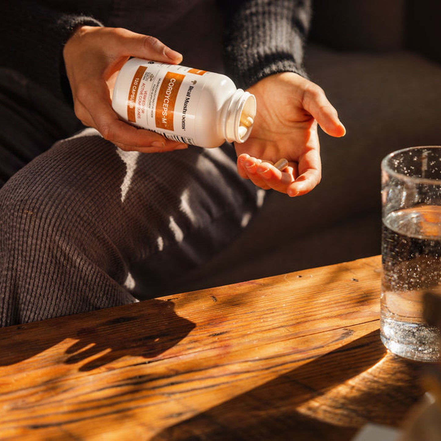 A person pours pills from a bottle of Organic Cordyceps Extract Capsules by Real Mushrooms into their hand over a wooden table, with a glass of water nearby. The label reads "Certified Organic" and highlights its inclusion of Cordyceps militaris mushrooms, renowned for their high beta-glucan content.