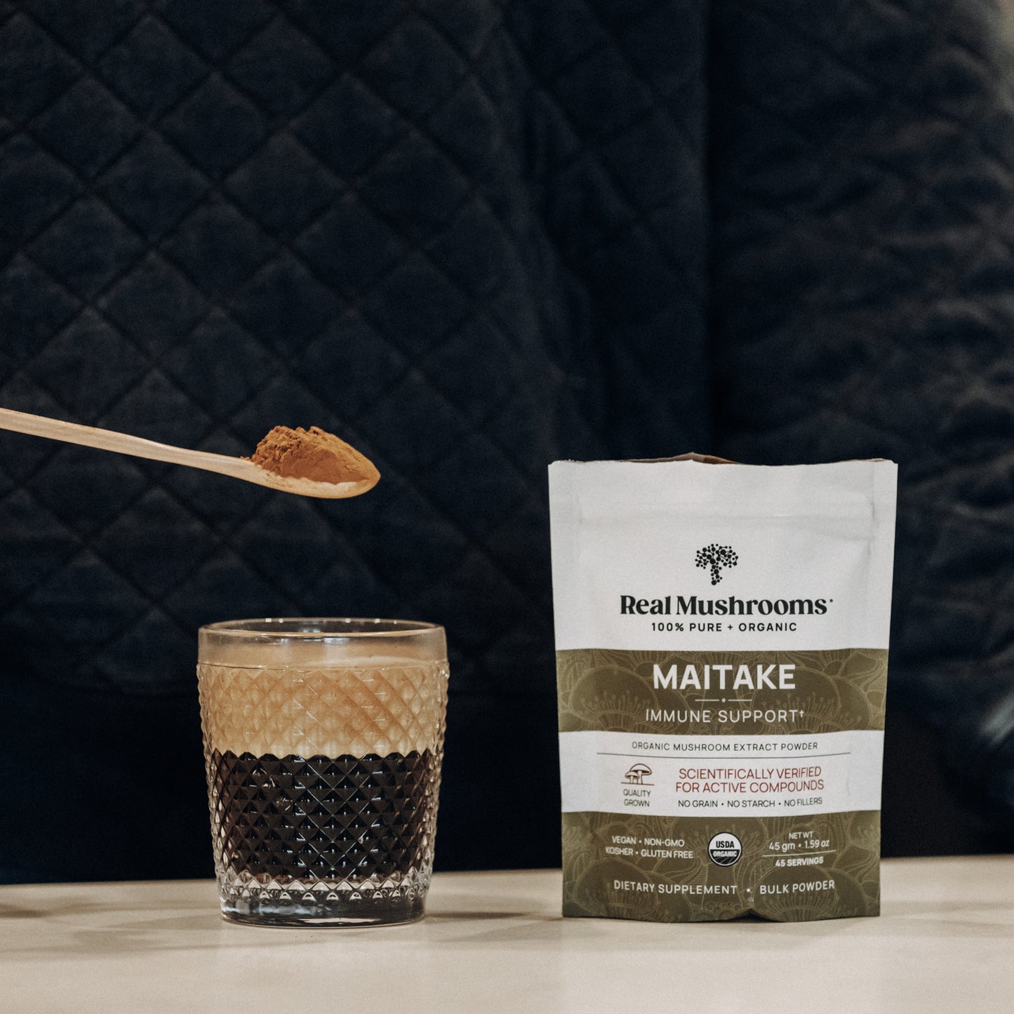 A person holds a spoon of Organic Maitake Mushroom Powder – Bulk Extract above a glass of coffee. A bag of Real Mushrooms' maitake powder, rich in beta-glucans, is on the table.