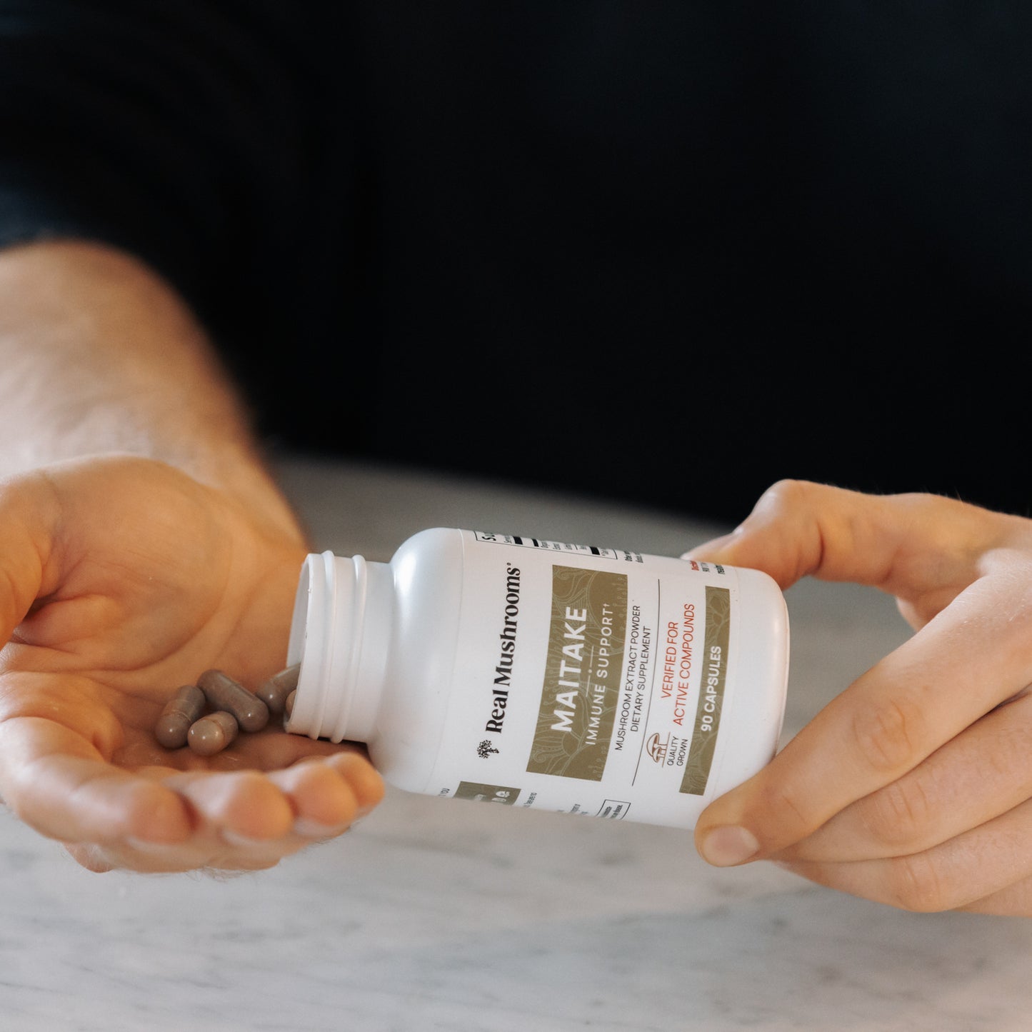 A person pours capsules from a white "Organic Maitake Mushroom Capsules" bottle by Real Mushrooms onto their hand, highlighting the benefits as vegan supplements rich in beta-glucans, known for supporting wellness and immune health.