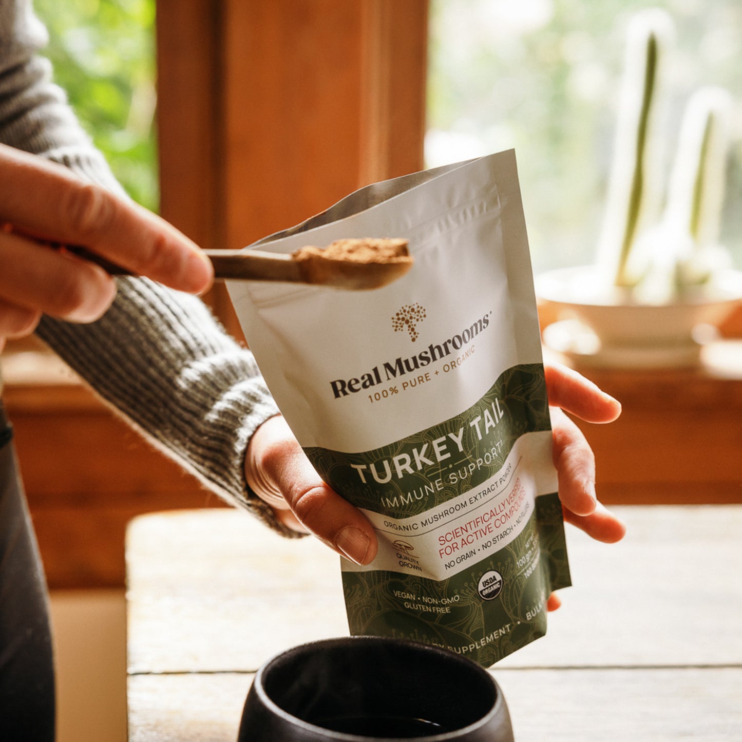 A person scoops some bulk powder from a bag of Turkey Tail Extract by Real Mushrooms into a bowl, with a window and plants visible in the background.