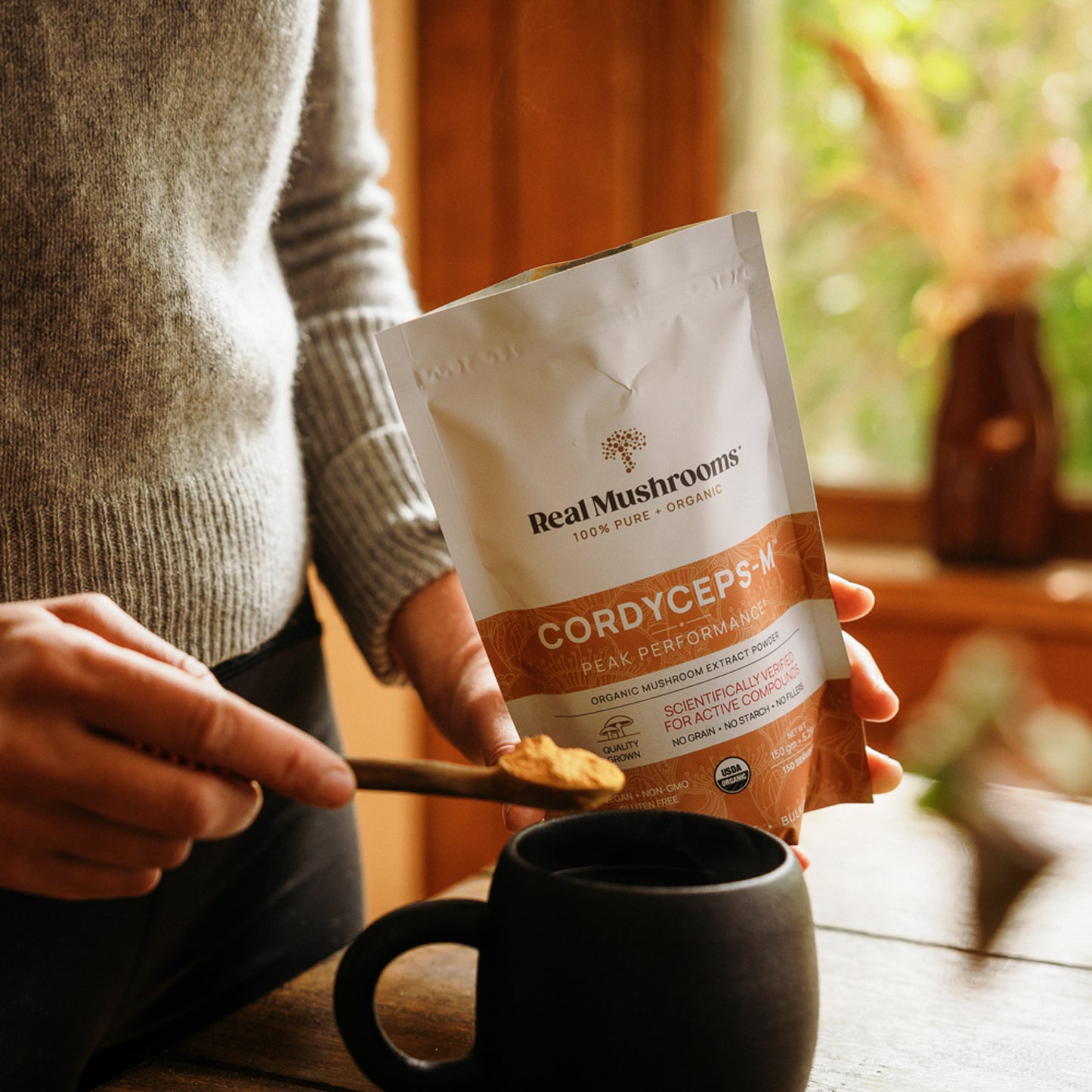 In a sunlit room, a person thoughtfully adds Real Mushrooms' Organic Cordyceps Mushroom Extract Powder – Bulk Supplement to a mug.