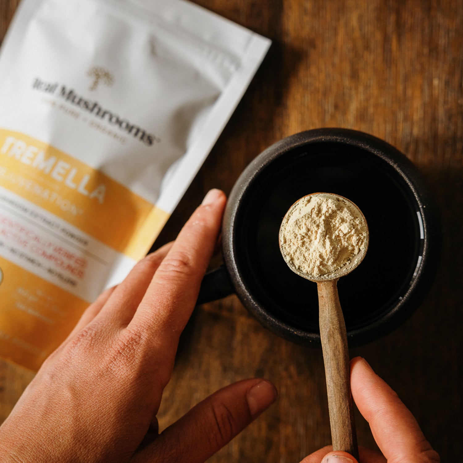 A hand holds a wooden spoon filled with the Organic Tremella Extract Powder above a black cup, suggesting the rich infusion of Tremella fuciformis mushrooms. Nearby, a packet labeled with Real Mushrooms rests on the wooden surface, proudly marked as USDA Certified Organic.