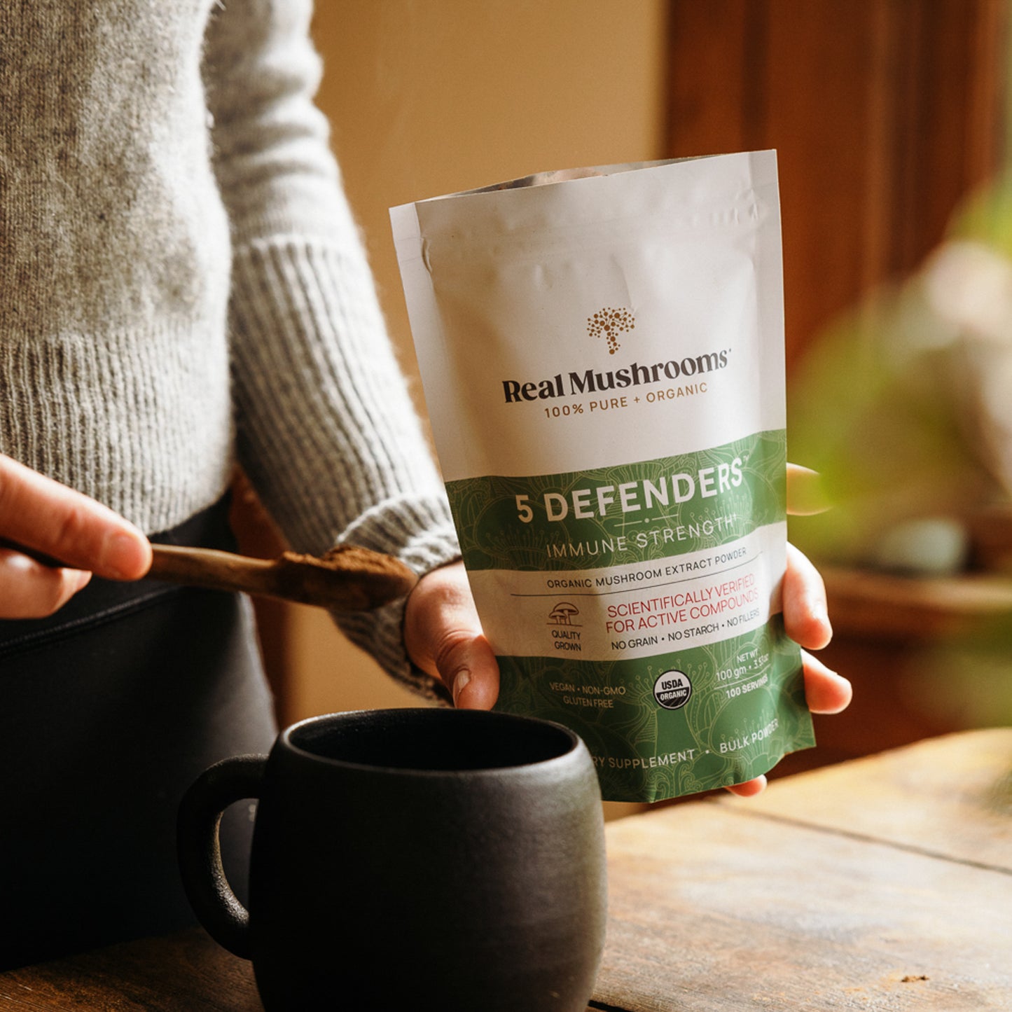 A person is holding a bag labeled "5 Defenders Organic Mushroom Complex – Bulk Powder" by Real Mushrooms and stirring its potent contents, rich in beta-glucans, into a black mug.